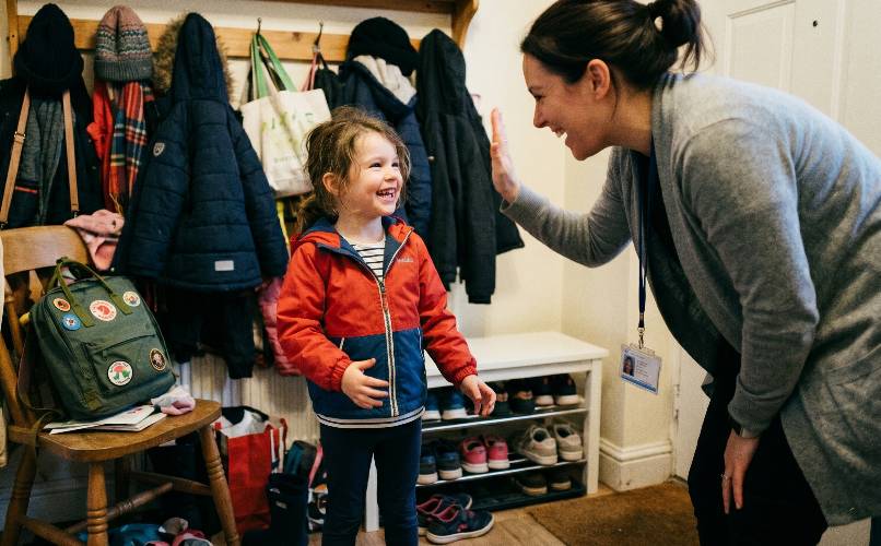 A child in a red jacket high-fiving a smiling specialist beside the coat hooks at home.