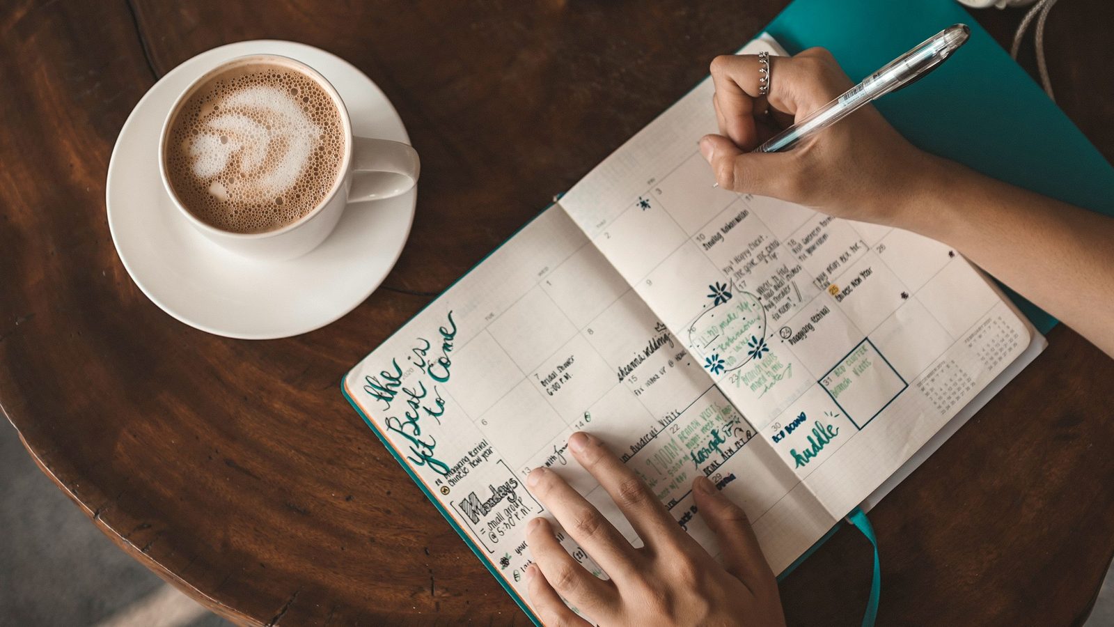 Hands fill in a monthly planner with a pen next to a cup of coffee on a dark wooden table.